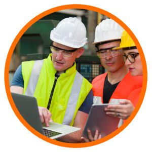 Factory workers in safety gear reviewing information on a laptop and tablet during a shop floor discussion.